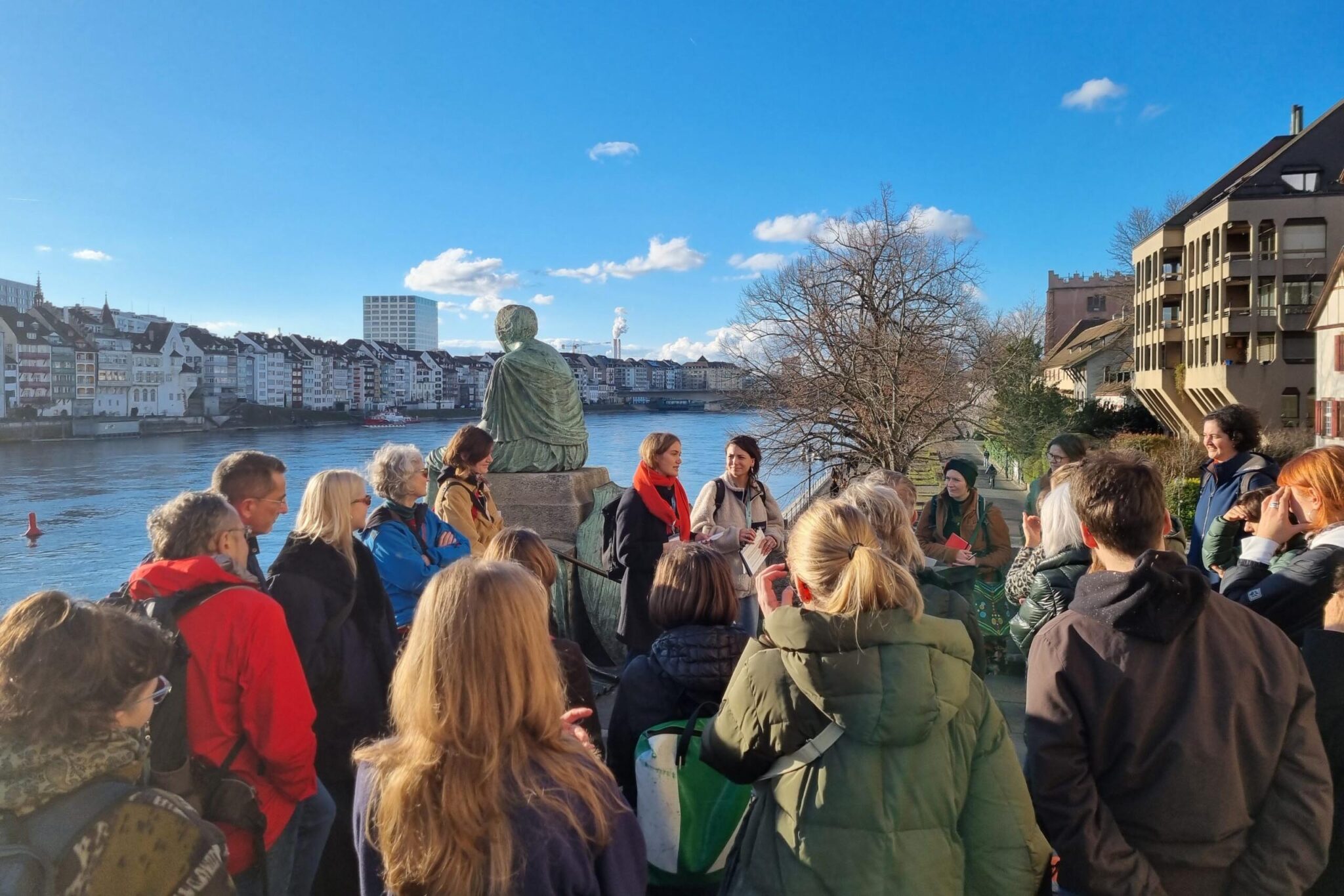 Zwei Stadtführerinnen begrüssen eine grosse Menge an Besucher:innen. Sie stehen zusammen vor der Helvetia-Statue auf der Mittleren Brücke. Hinter ihnen der Rhein und blauer Himmel.