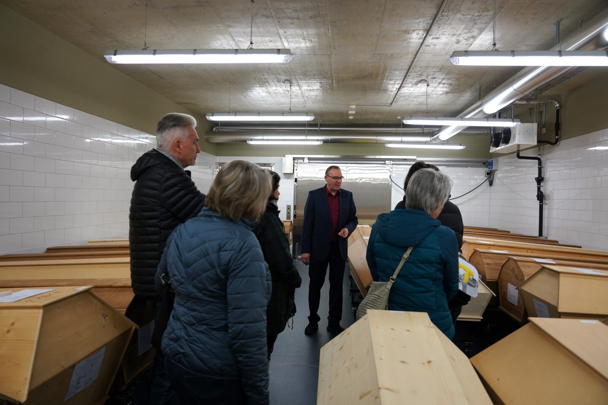 In mitten von geschlossenen Holzsärgen stehen fünf Menschen in dicken Jacken und ein Mann in Hemd und Jacket. Er trägt eine Brille und spricht. An der Decke hängen Neonröhren und Lüftungsröhren und die Wände sind weiss geplättelt.