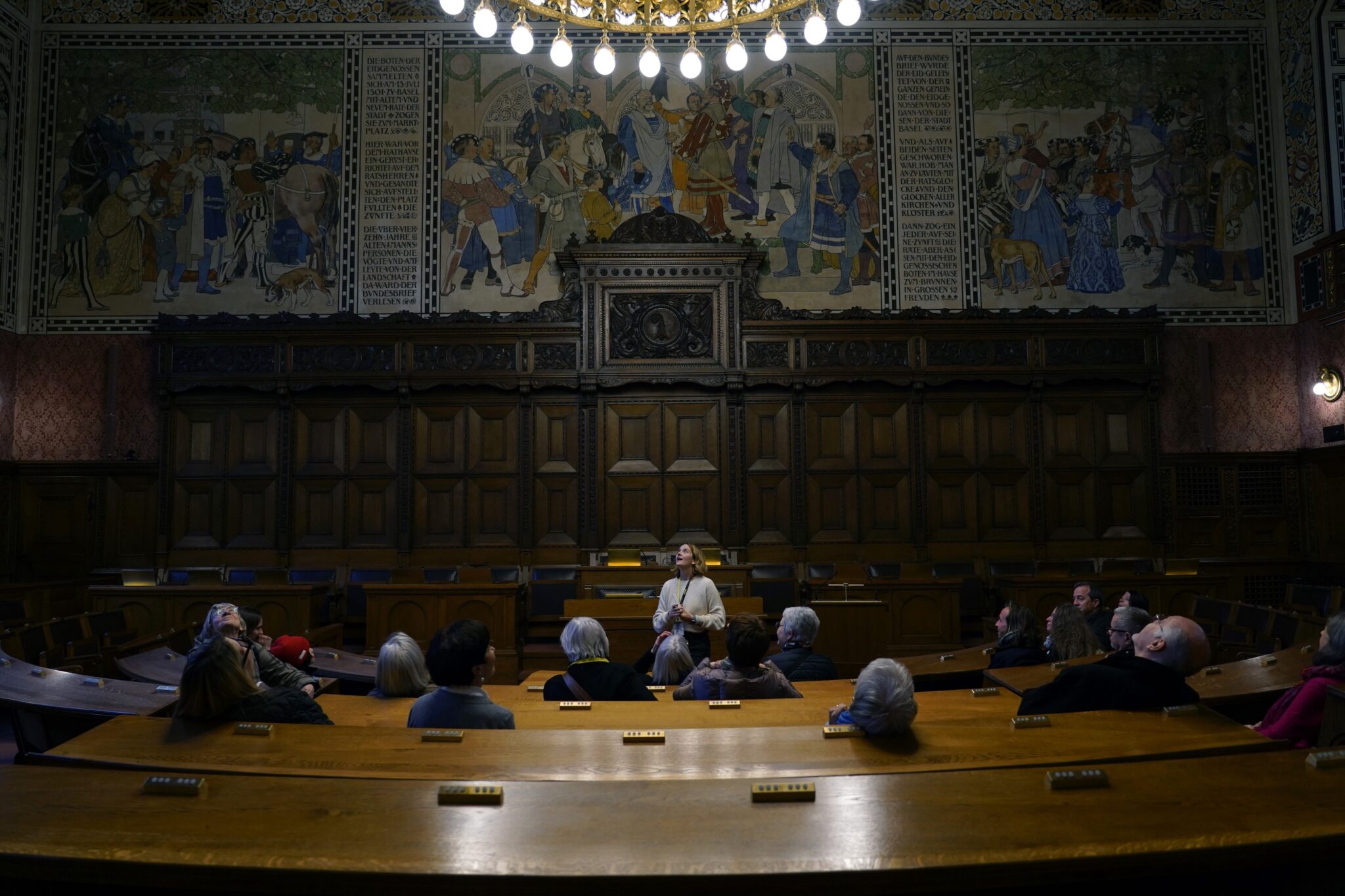 Der Grossratssaal im Basler Rathaus. Von der Decke scheint ein riesiger Kronleuchter. Darunter sitzen in den Holzreihen Zuschauende. Sie blicken nach vorne und nach oben an die Decke. Die Wände sind zur einen Hälfte mit dunklem Holz verkleidet und zur anderen mit Bildern bemalt.