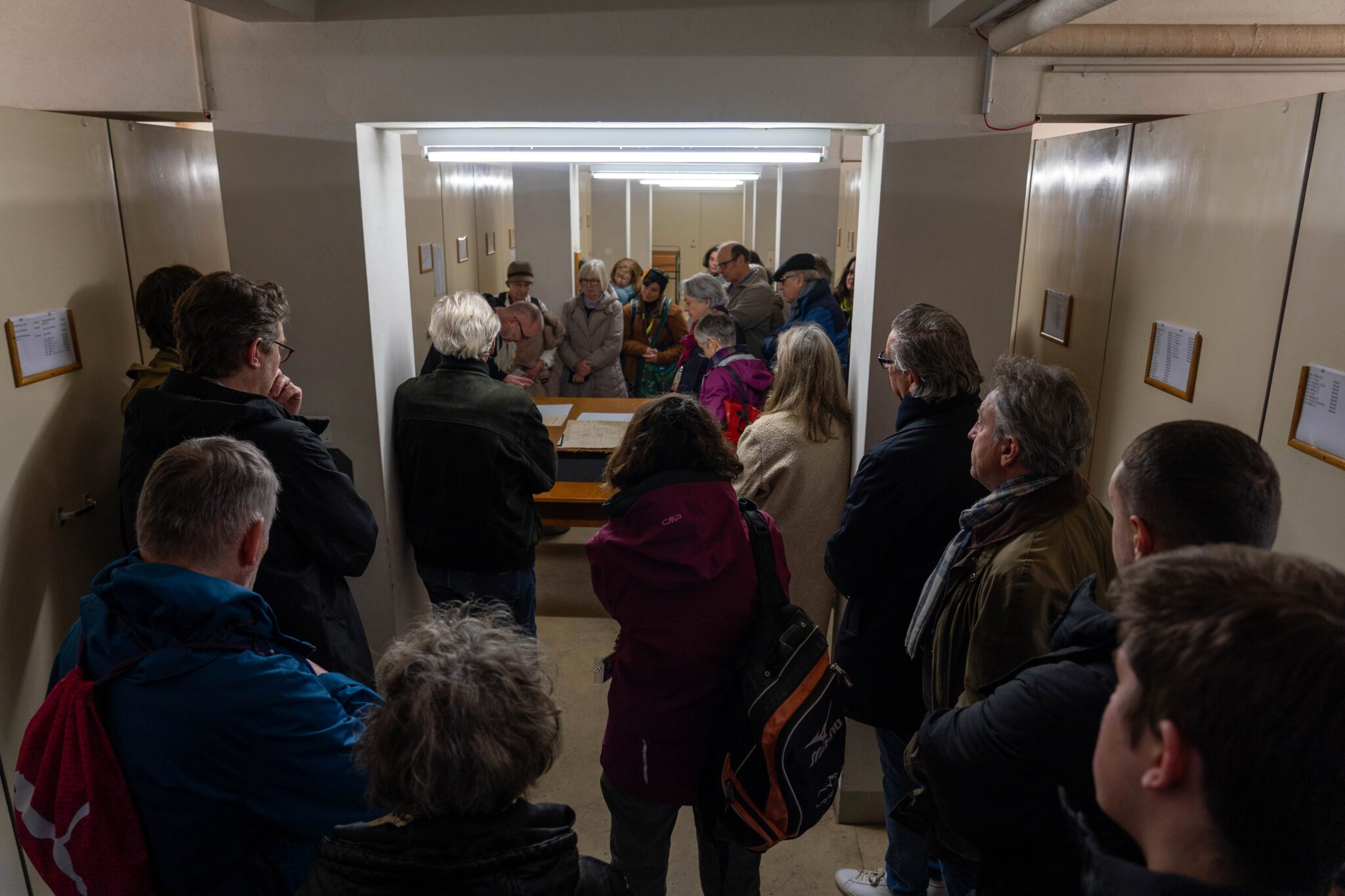 Menschen versammeln sich in einem Aktenlager um einen Tisch. Links und rechts von ihnen stehen geschlossene grau-beige Aktenschränke.