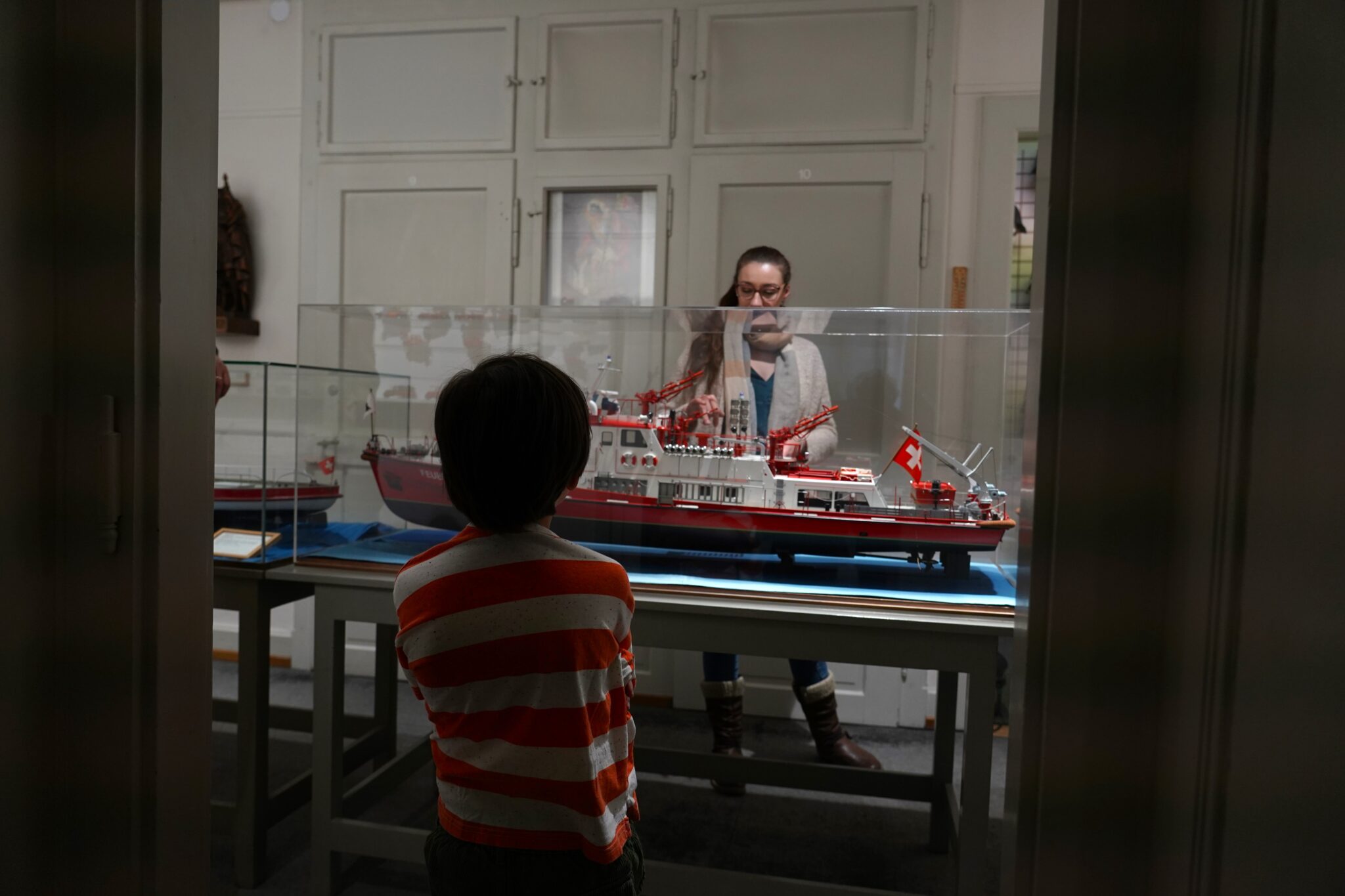 Ein Kind in rot-weiss gestreiftem T-Shirt blickt auf ein riesiges Modell-Schiff in einer Vitrine. Dahinter steht eine Frau und zeigt erklärend auf das Schiff. Ihr Gesicht ist bis zur Hälfte vom Glas der Vitrine verdeckt.