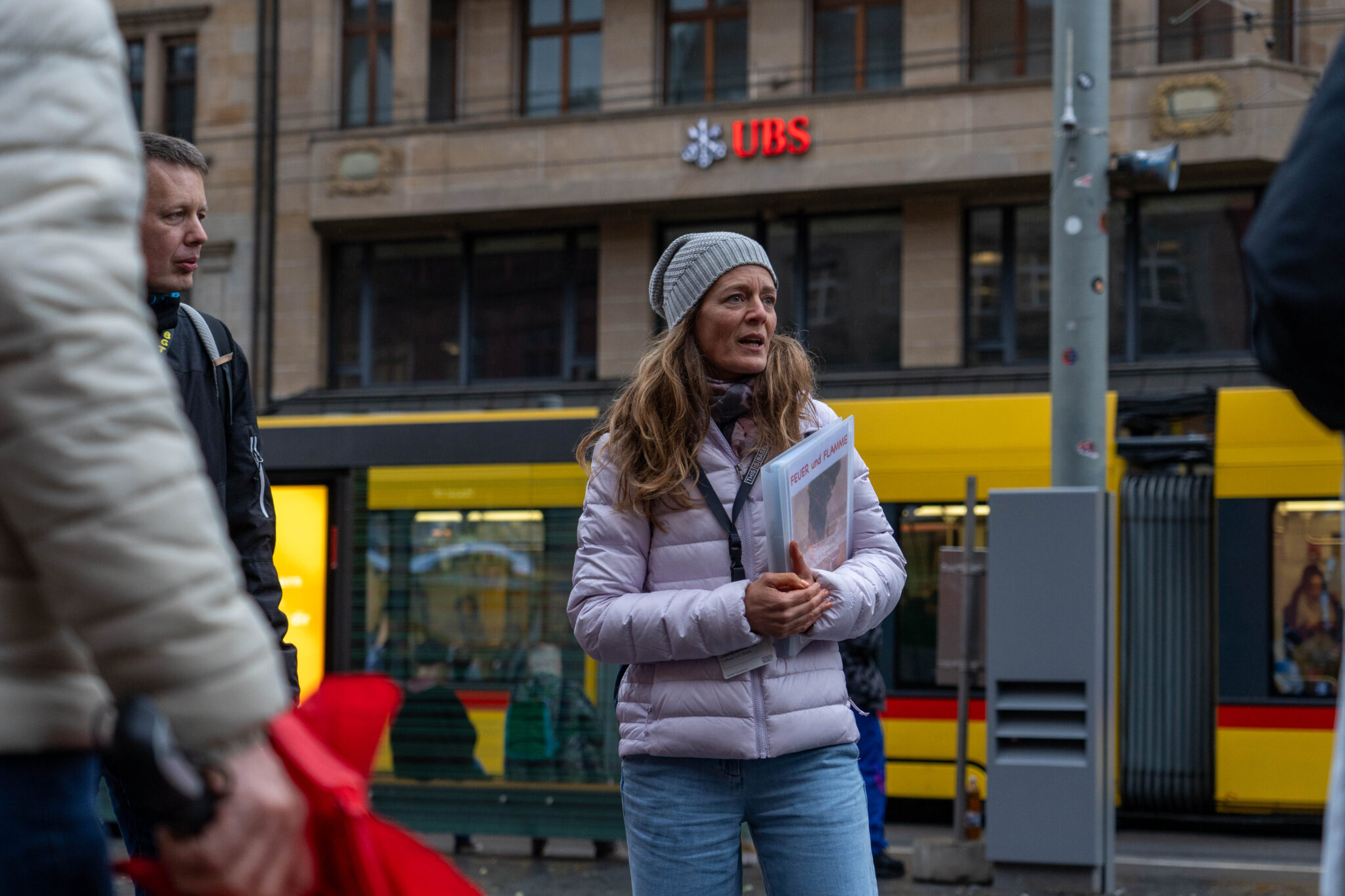 Eine Stadtführerin in rosa Daunenjacke, grauer Mütze und blauen Jeans erzählt etwas. In den Händen hält sie eine Mappe mit Aufschrift: Feuer und Flamme. Im Hintergrund fährt ein gelbes Tram vorbei.