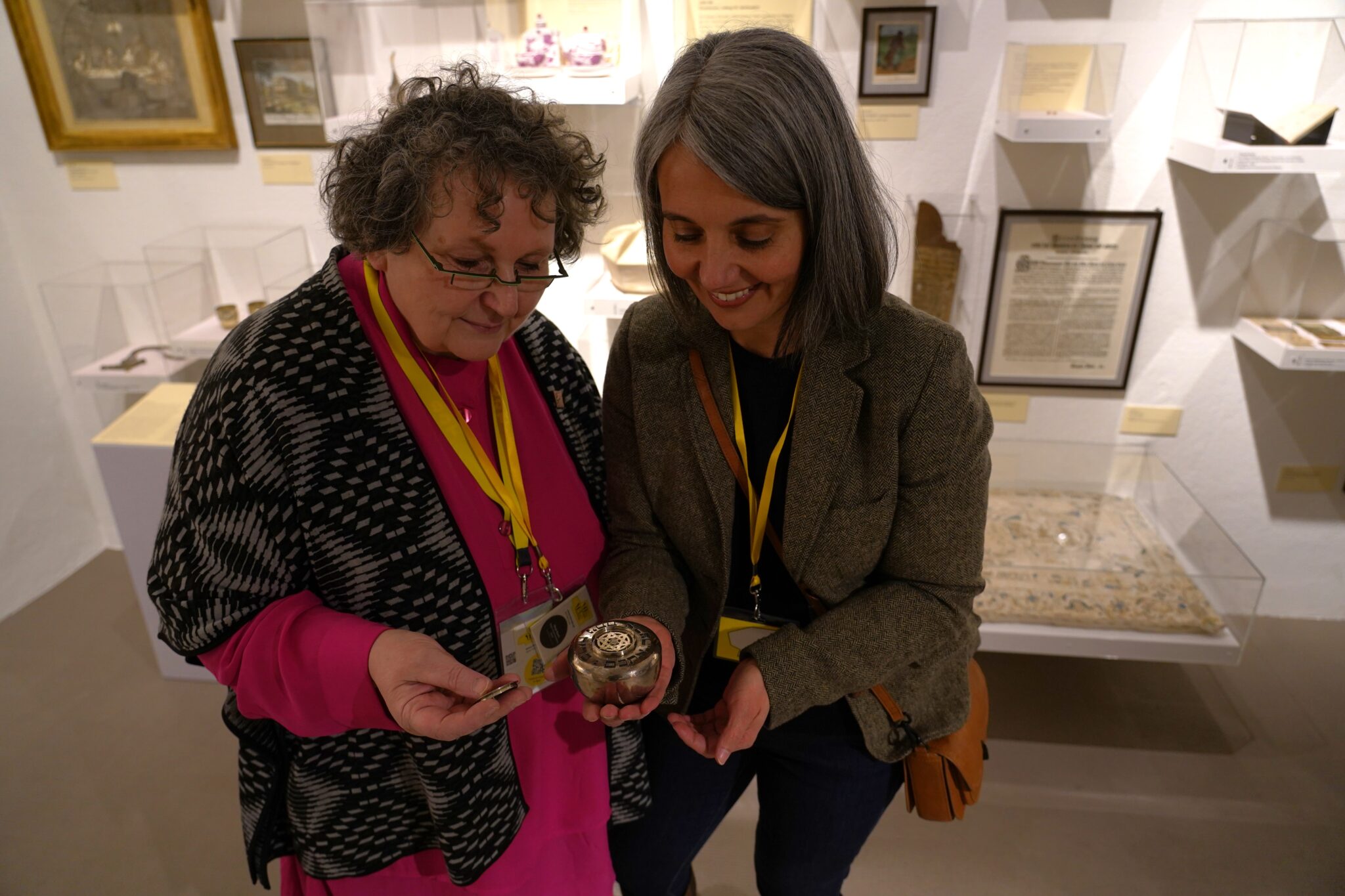 Zwei weiblich gelesene Frauen halten Gegenstände der jüdischen Kultur in den Händen. Die eine trägt eine Brille und ein pinkes Kleid, die andere eine graue Jacke und eine braune Ledertasche. Sie stehen im Jüdischen Museum.