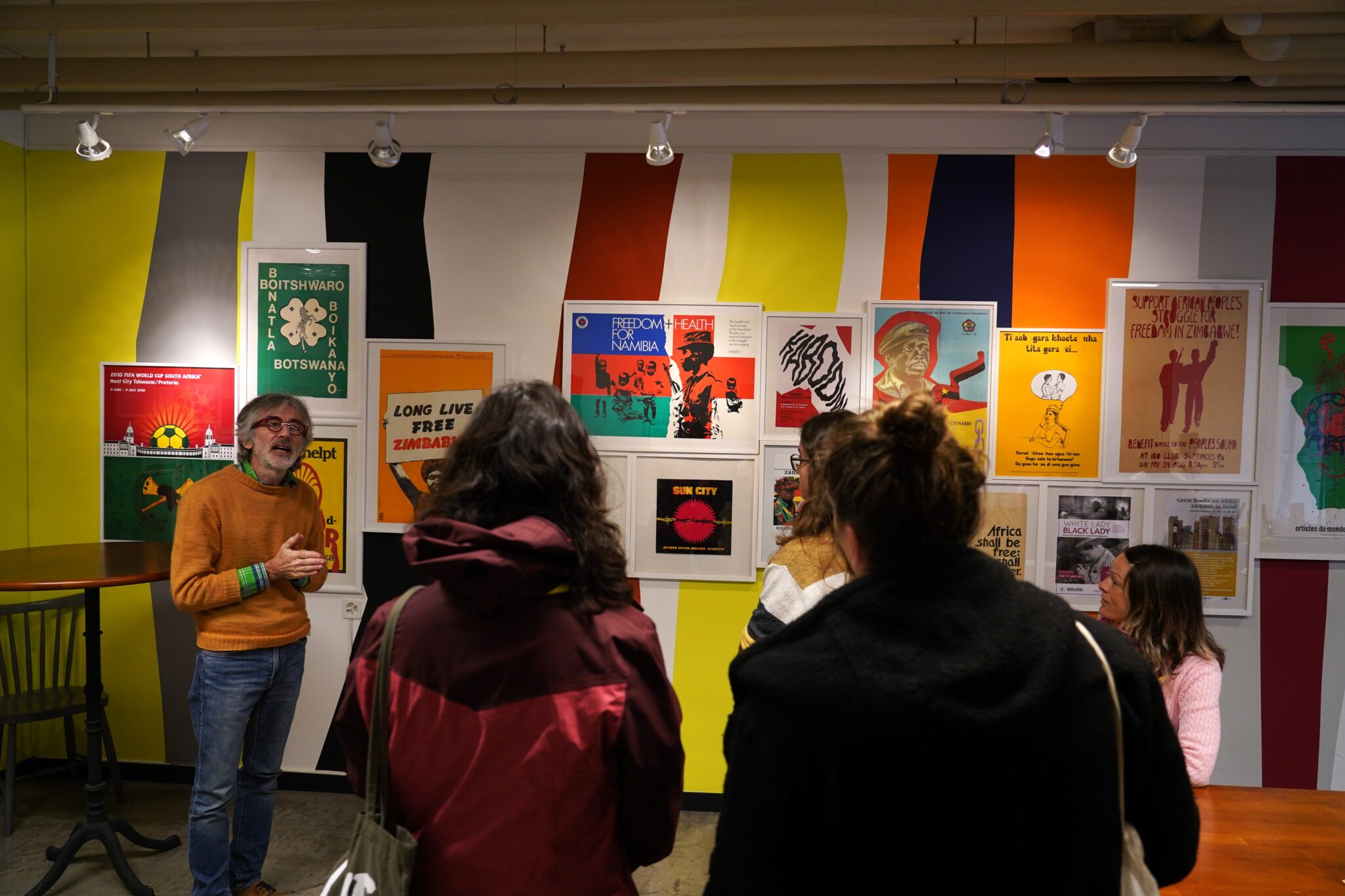 Ein Mann trägt einen orangenen Wollpullover, blaue Jeans und eine rote Brille. Er steht vor einer Plakatwand und erklärt einer kleinen Gruppe von Menschen etwas. Die Gruppe sieht man von hinten.