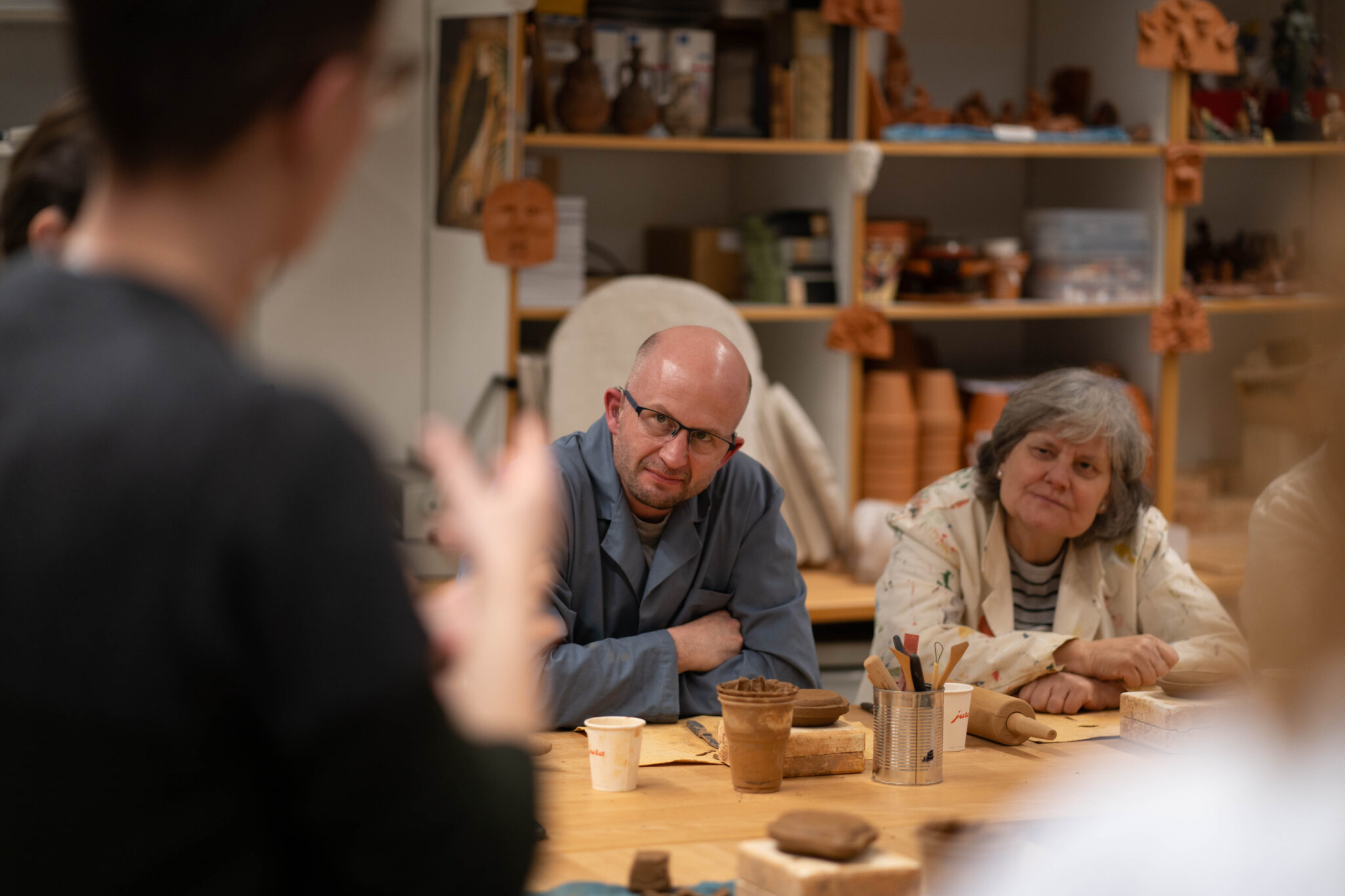 Zwei Personen sitzen an einem Tisch. Vor ihnen liegen ein Wallholz, ein Tonklumpen und Werkzeuge zum Ton modellieren. Sie blicken aufmerksam zu einer Person, die Gebärdensprache spricht.
