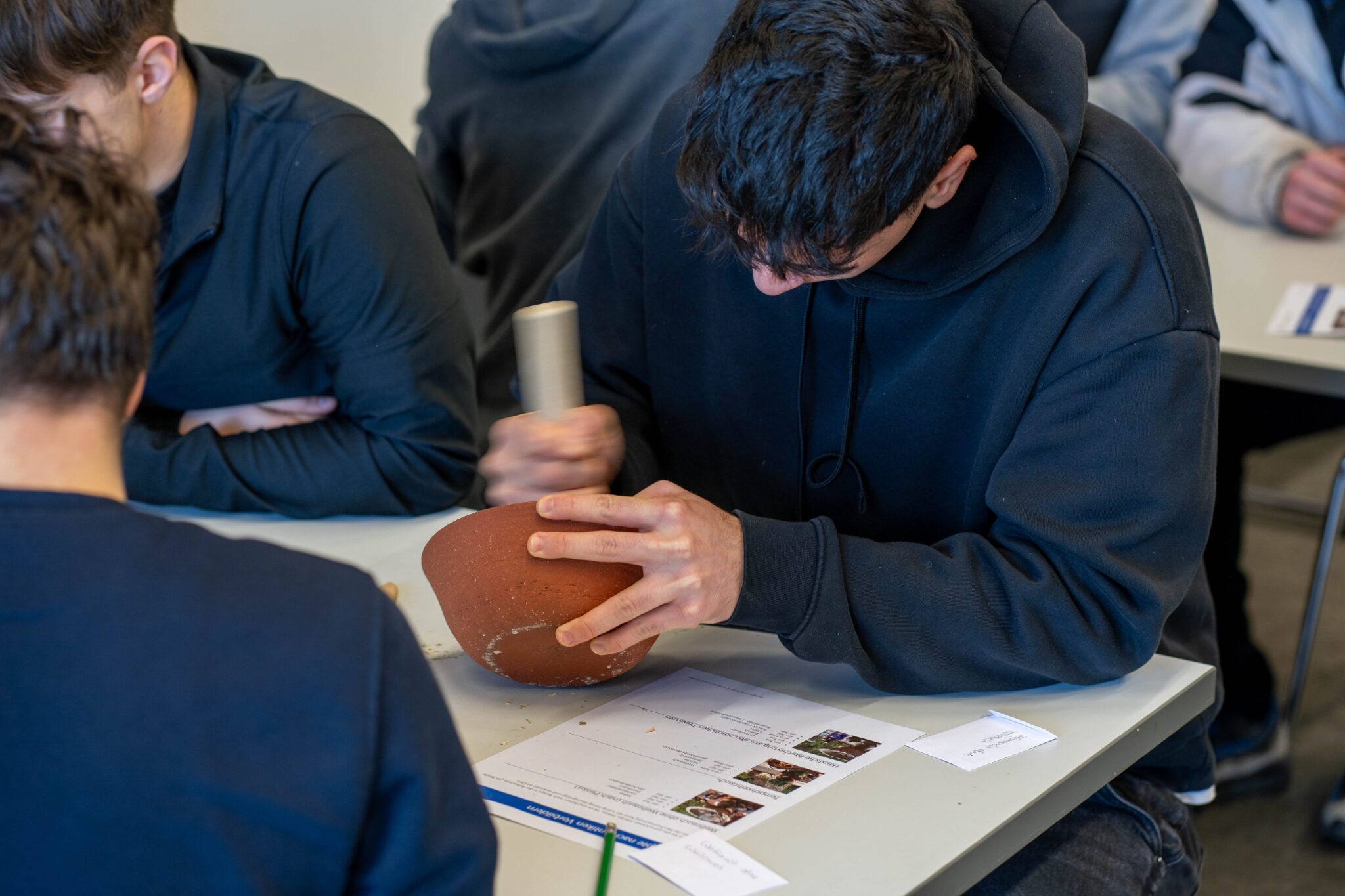 Ein Teenager in schwarzem Hoodie mörsert mit Schwung eine Räuchermischung in einer braunen Tonschale. Dabei sitz er an einem Tisch, neben ihm sitzen andere Schüler. Auf dem Tisch liegt ein Arbeitsblatt.