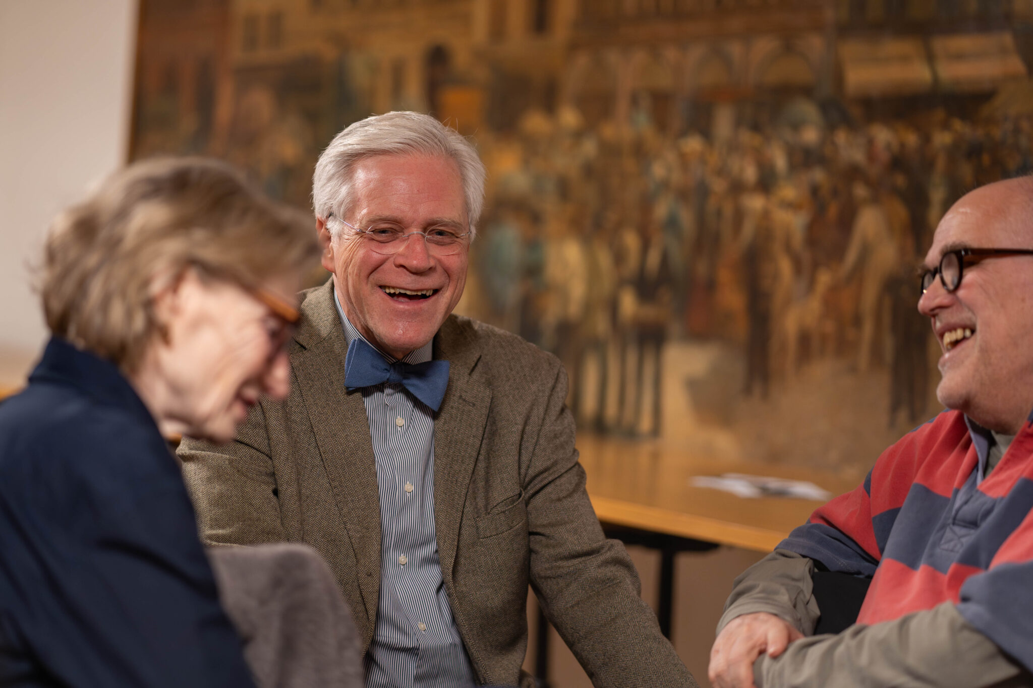 Drei Personen sitzen vor einem meterhohen Gemälde. Sie tragen alle eine Brille und lachen herzlich.
