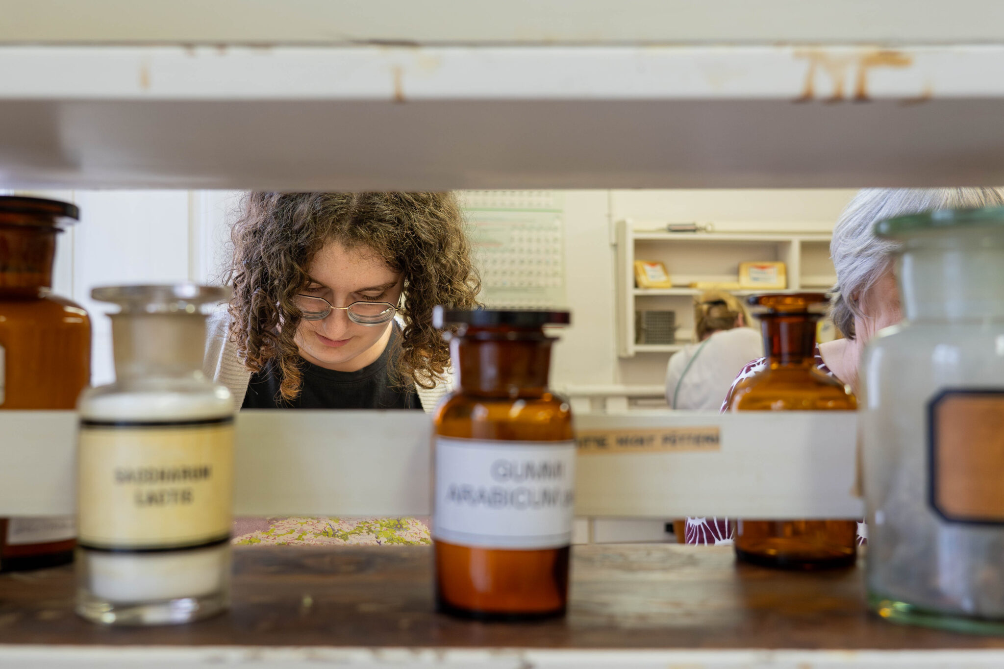 Auf einem Regal stehen alte medizinische Flaschen mit Glasdeckeln. Dahinter erkennt man die Köpfe von einer jungen und einer älteren Person. Sie blicken nach unten.