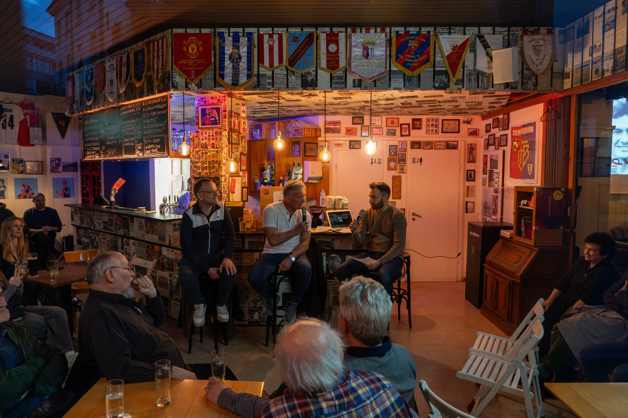 Drei Männer sitzen auf Barhockern und sprechen in ein Mikrophon. Sie sind in einer Fussballbar. An den Wänden hängen Paninibilder, Fotografien und Fussballfanartikel. Es ist etwas düster. Nur 5 Glühbirnen erhellen den Raum.