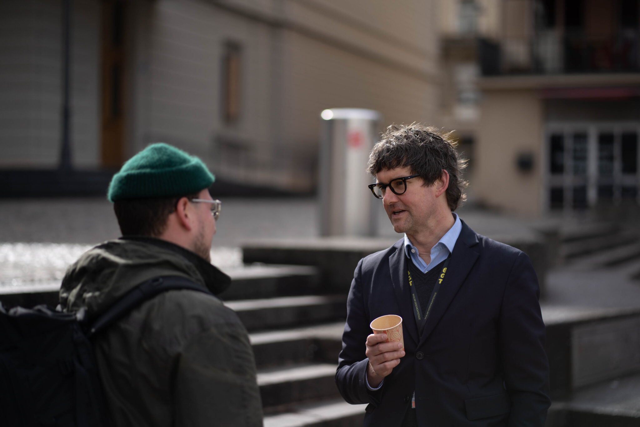 Zwei Personen unterhalten sich. Beide sind dunkel gekleidet. Der eine trägt eine grüne Mütze, der andere einen Kaffeebecher in der Hand.