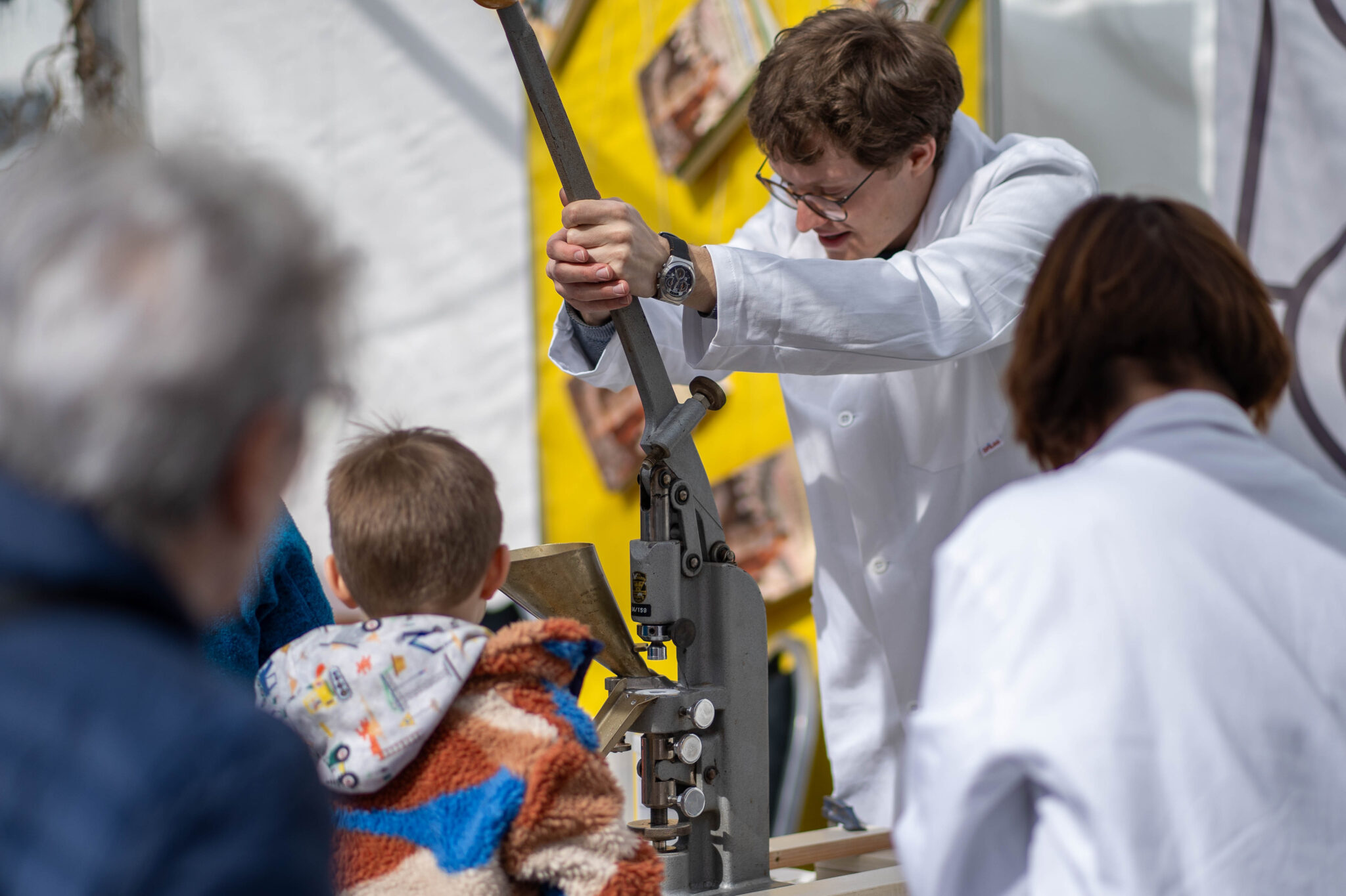 Ein junger Mann in einem weissen Kittel zieht am langen Hebel einer alten Tablettenpresse. Ein Kind schaut ihm dabei zu.