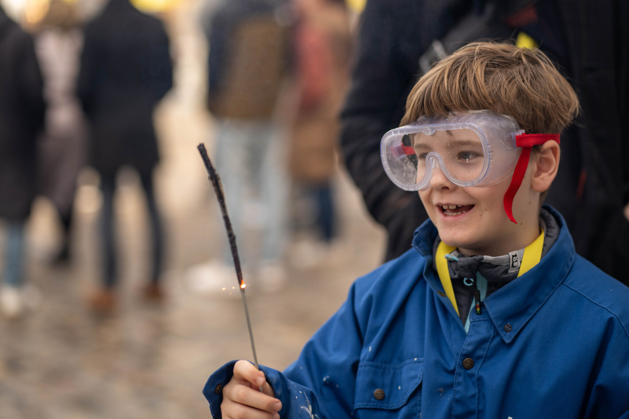 Ein Kind trägt eine grosse durchsichtige Schutzbrille mit einem roten Bändel und ein blaues Arbeitsgewand. In der Hand hält das Kind eine brennende Wunderkerze.