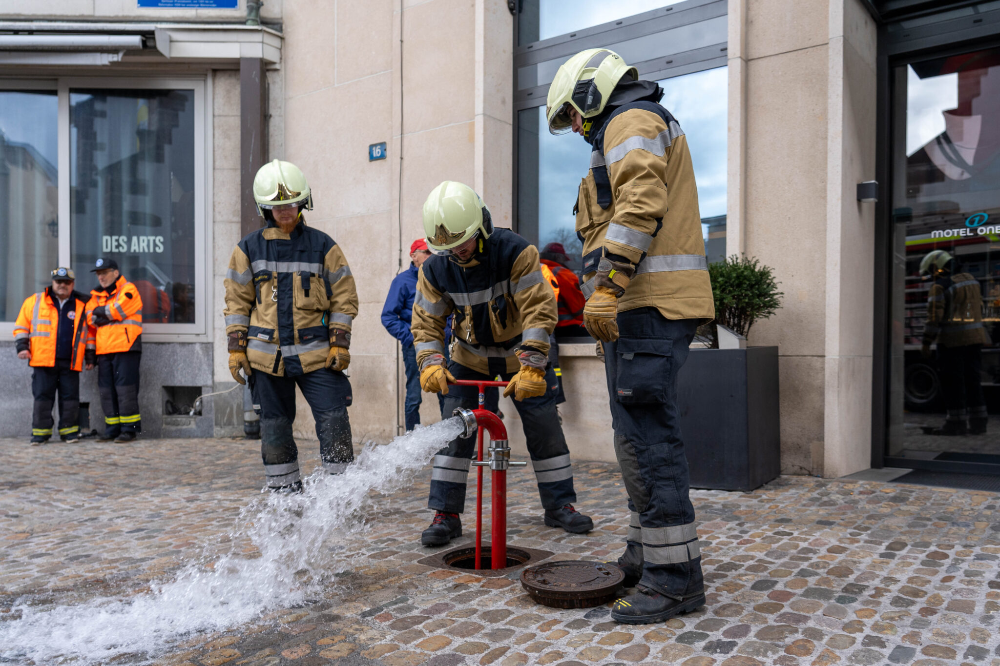 Drei Feuerwehrmänner mit Helm und Handschuhen lassen Wasser aus einem Hydranten.