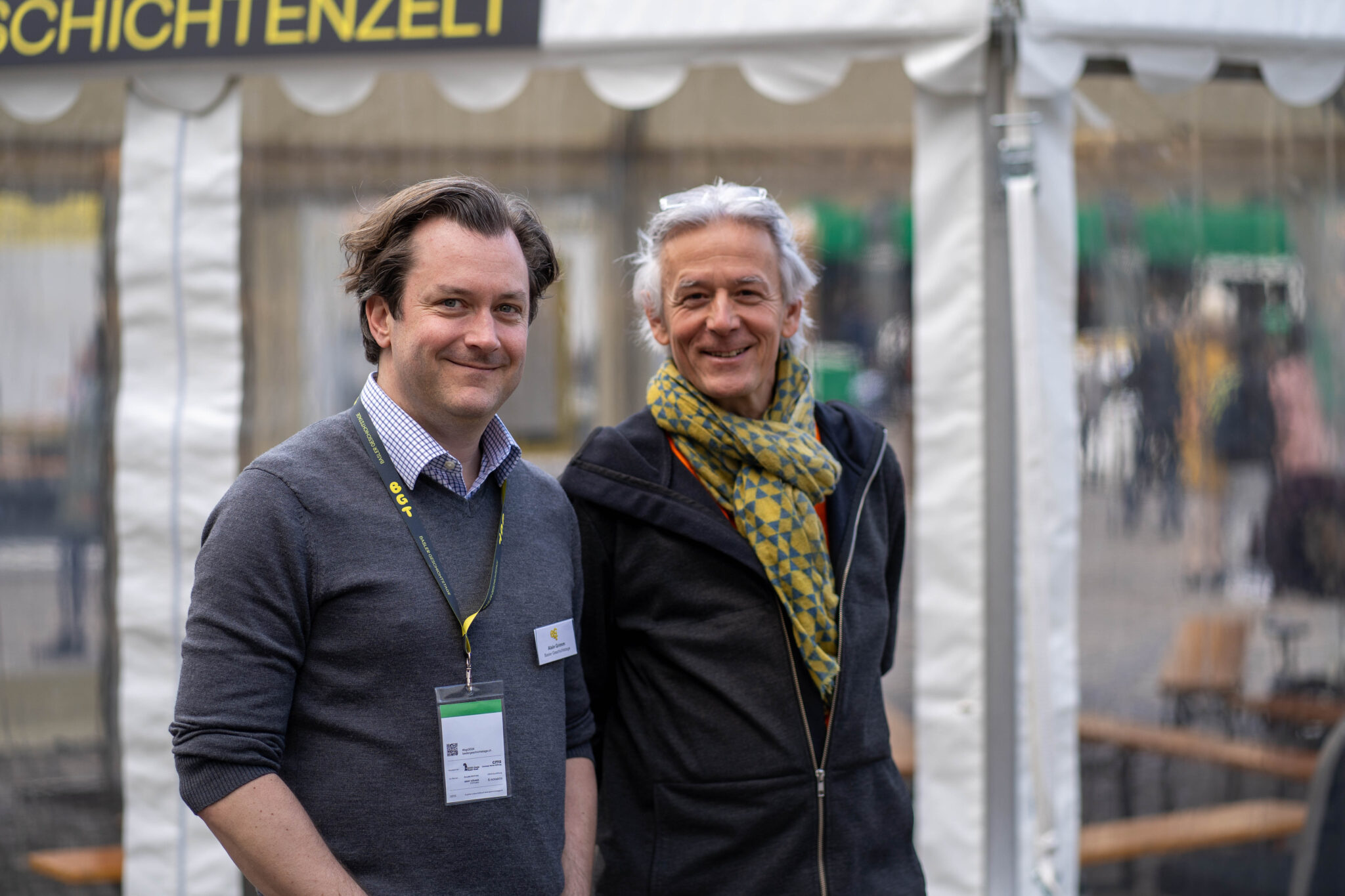 Zwei Männer stehen zusammen und lächeln in die Kamera. Beide tragen dunkle Pullover.
