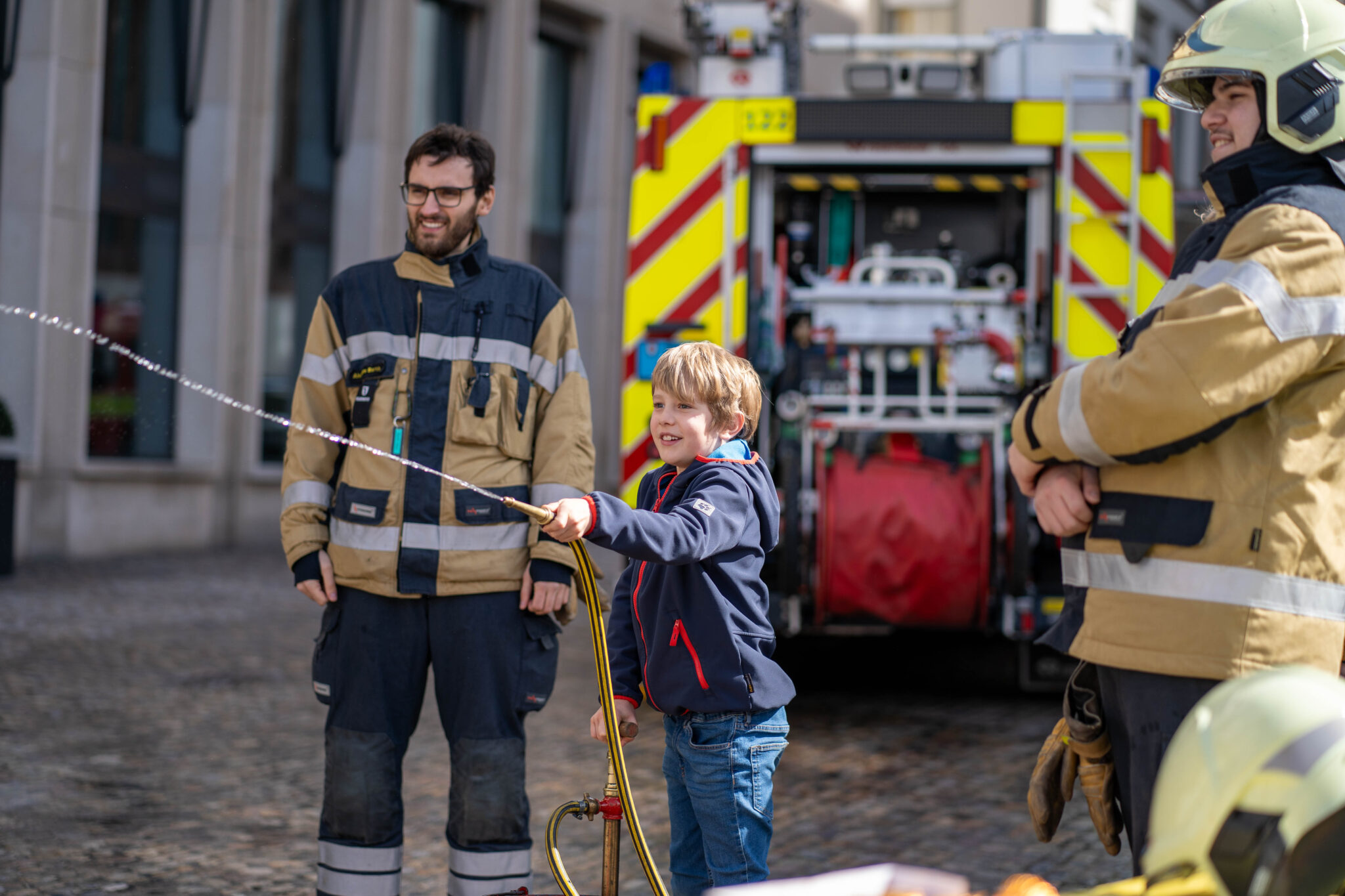 Zwei Feuerwehrmänner schauen einem Kind zu, wie es mit einem Schlauch Wasser spritzt. Im Hintergrund steht ein grosses Feuerwehrauto.