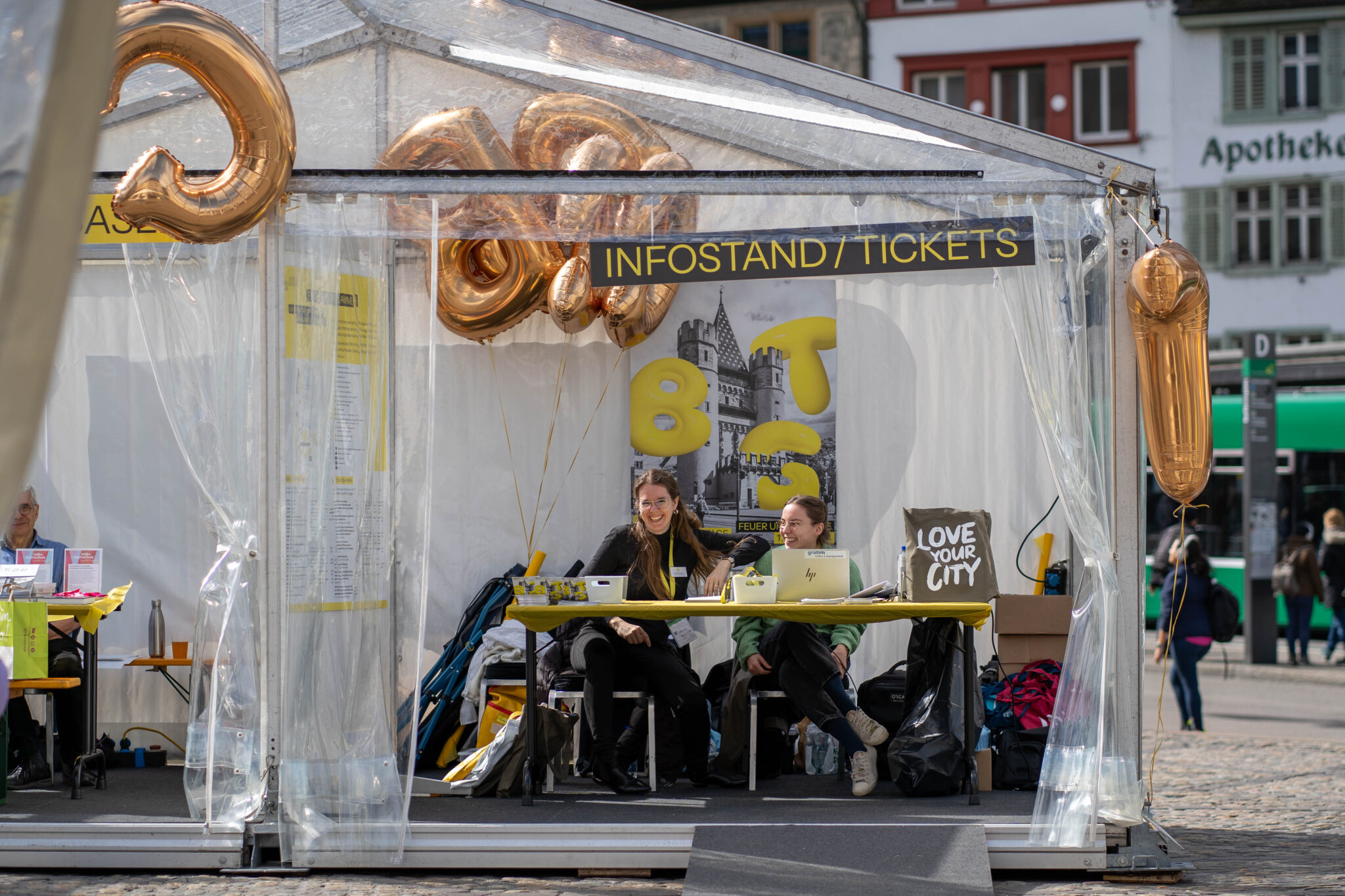 Ein transparentes Zelt mit Beschriftung: Infostand/Tickets. Darin steht ein Tisch, an dem zwei Personen sitzen und in die Kamera lachen.