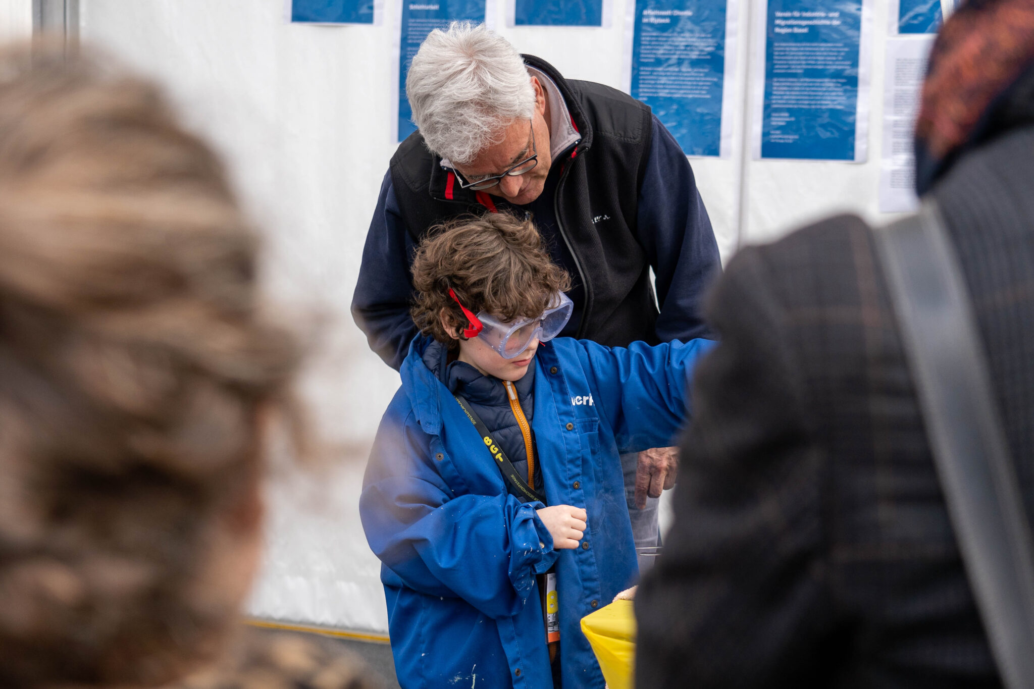 Ein Kind trägt eine grosse durchsichtige Schutzbrille mit einem roten Bändel und ein blaues Arbeitsgewand. Hinter ihm steht ein Erwachsener mit grauen Haaren und bäugt sich über die Schulter des Kindes.