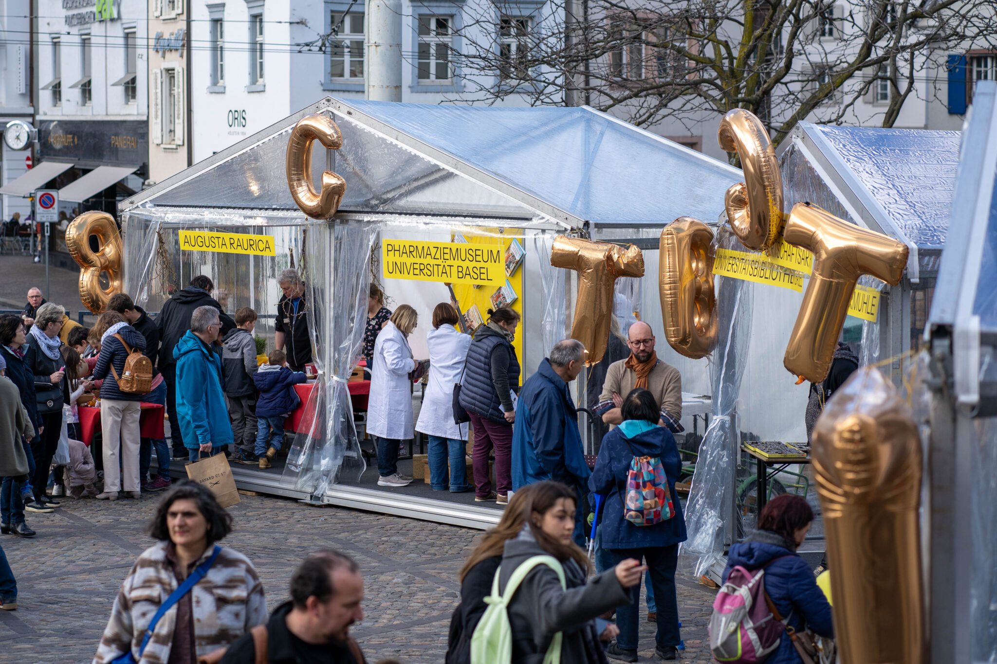 Zwei transparente Zelte. Aussen hängen goldene Ballone in Form von Buchstaben. Leute stehen vor den Zelten und sprechen zusammen.