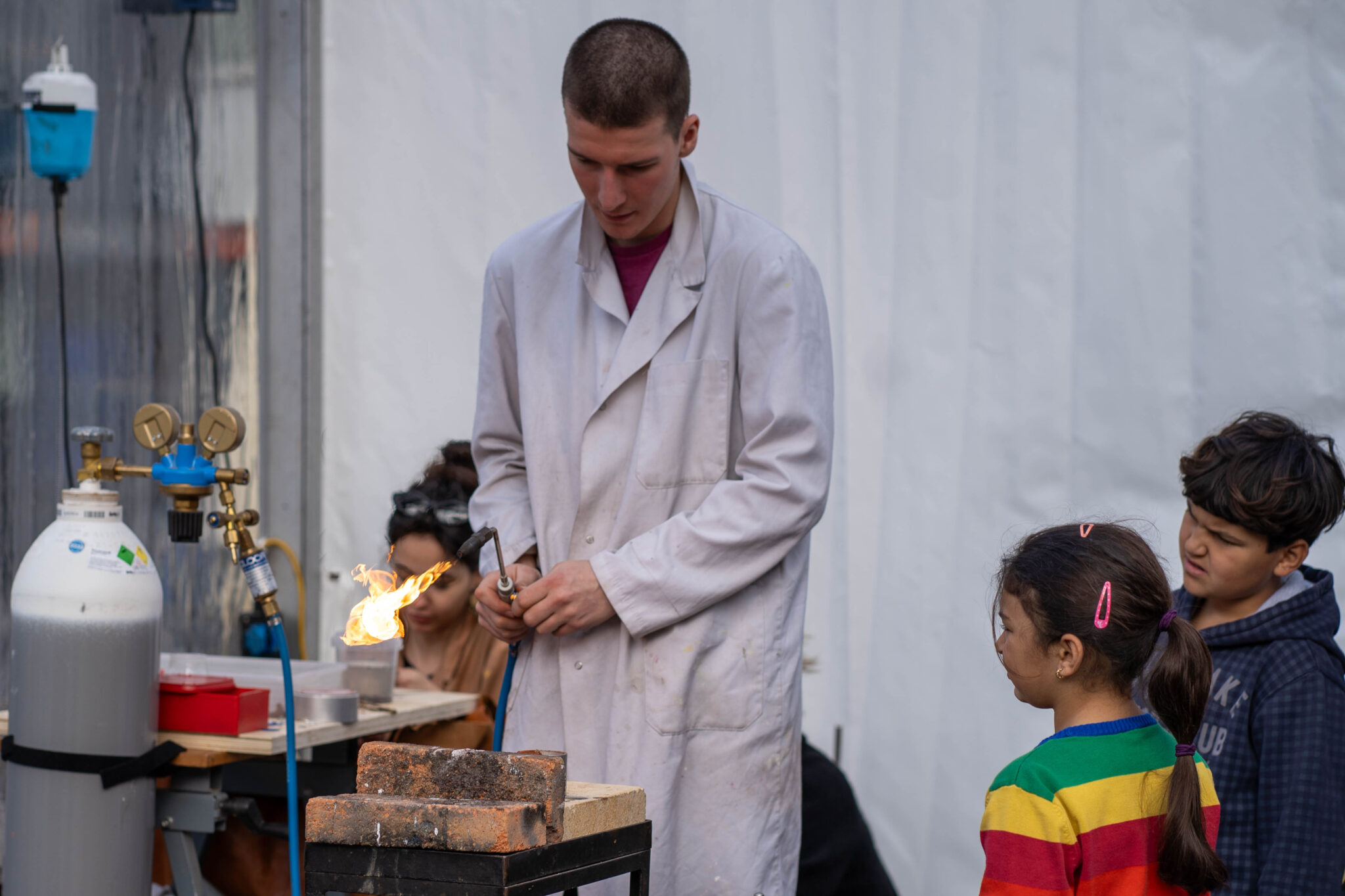 Ein junger Mensch mit weissem Arbeitsgewand bläst mit dem Bunsenbrenner eine Flamme in die Luft. Zwei Kinder schauen ihm dabei zu.
