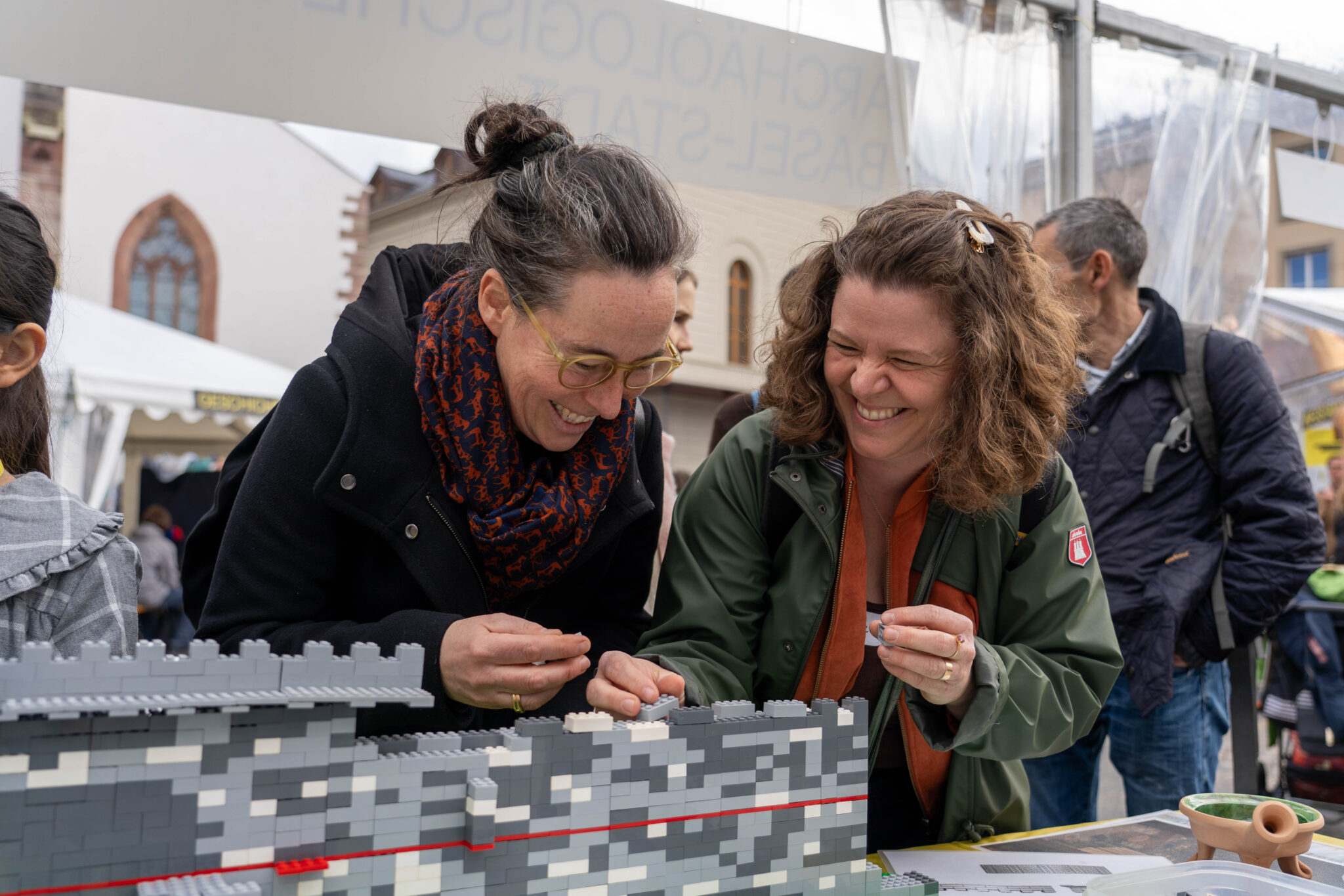 Zwei Menschen beugen sich über eine Mauer aus grauen Legosteinen und lachen. In den Händen halten sie Bausteine. Sie tragen Jacken und Schals.
