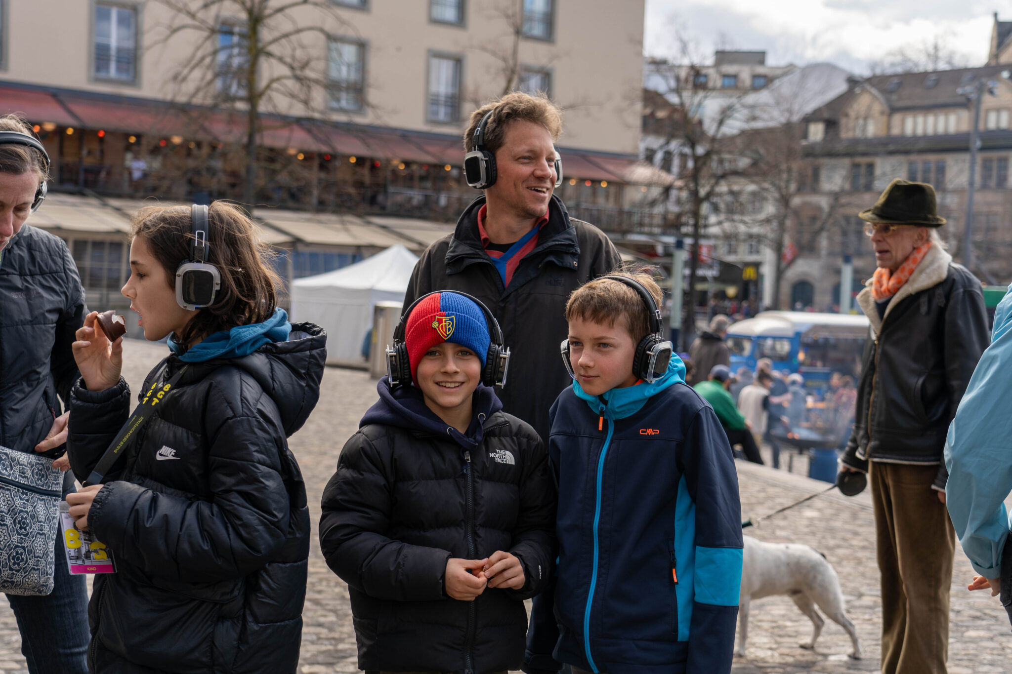 Besucher:innen des Fussballspaziergangs lachen in die Kamera. Sie tragen alle Kopfhörer. Das eine Kind trägt eine Mütze in den Farben des FC Basels.