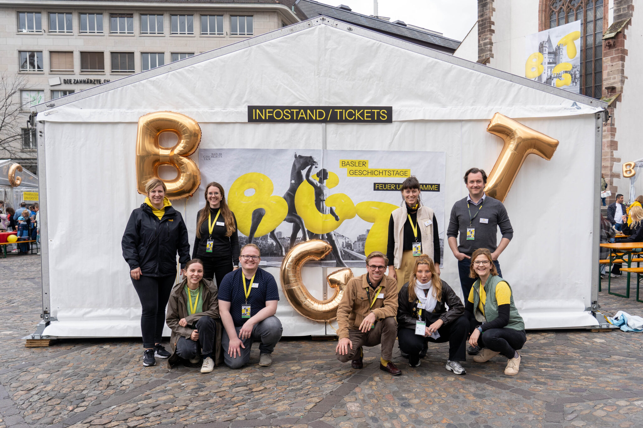 Neun Personen posieren vor einem Plakat der Basler Geschichtstage. Sie tragen alle einen gelben Bändel und lachen.